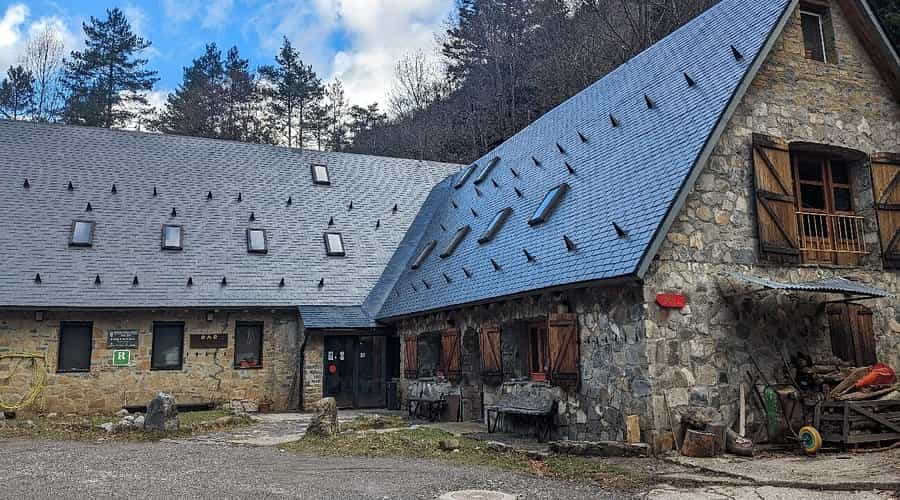 Stone refuge building with dark slate roof and wooden shutters in a forested mountain setting.