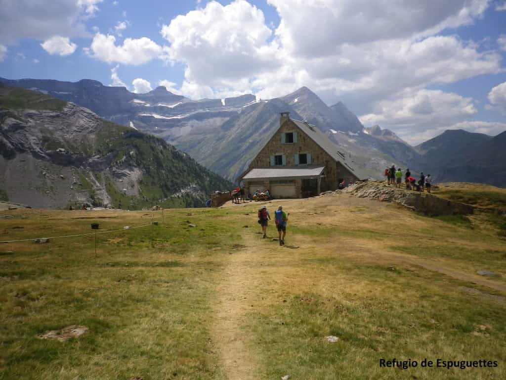 Hikers approaching Refugio de Espuguettes with snow-capped mountains under a cloudy sky.