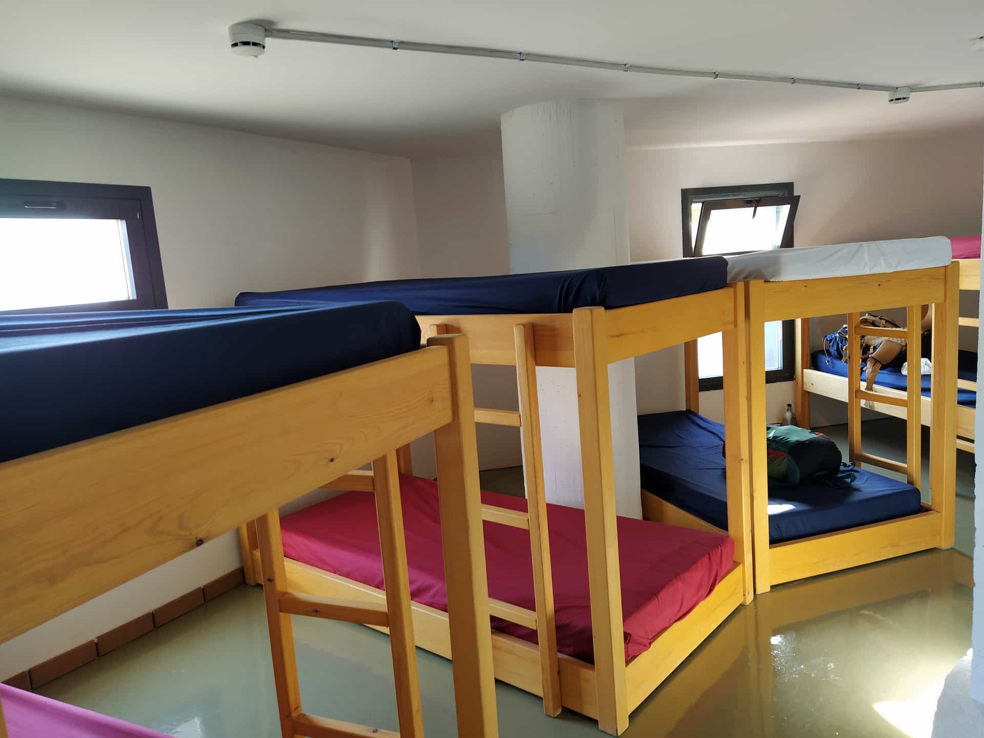 Wooden bunk beds with blue and red mattresses inside a bright dormitory room, likely a refuge.