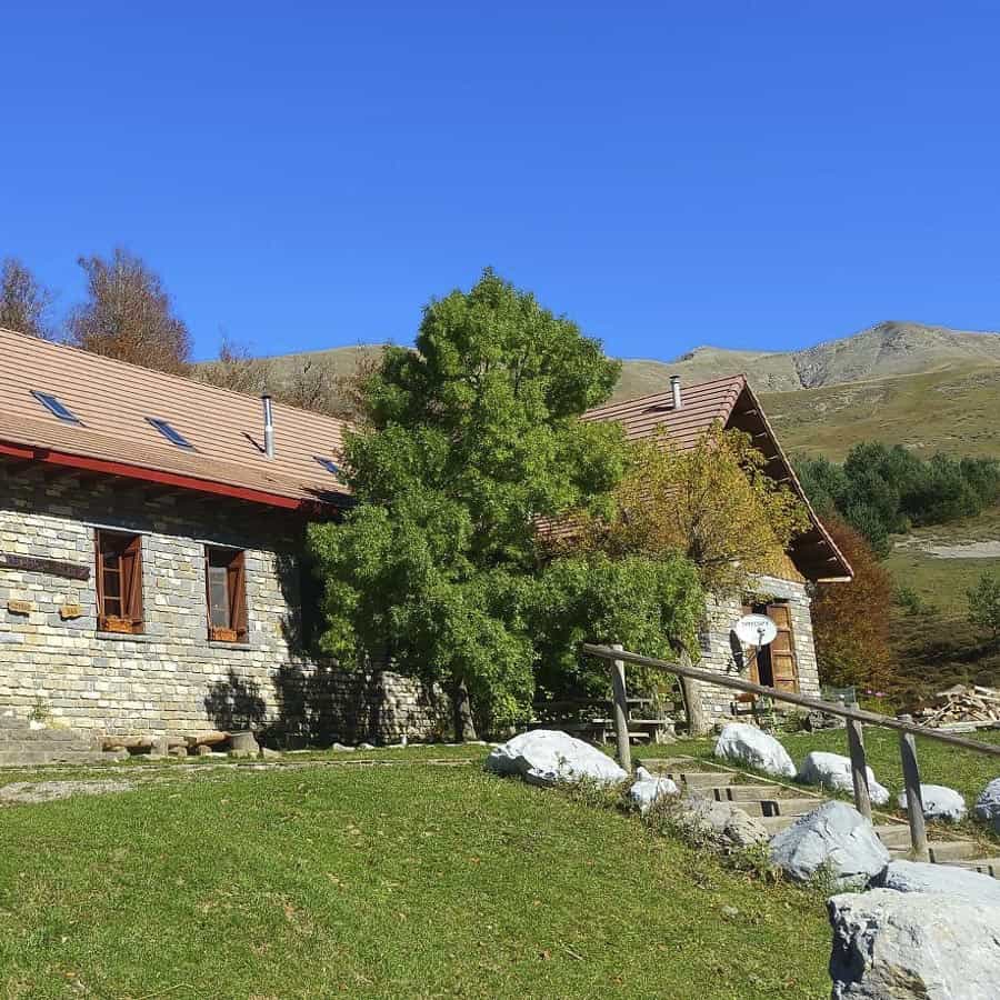 Stone refuge building with green lawn and mountains under clear blue sky, Linza.
