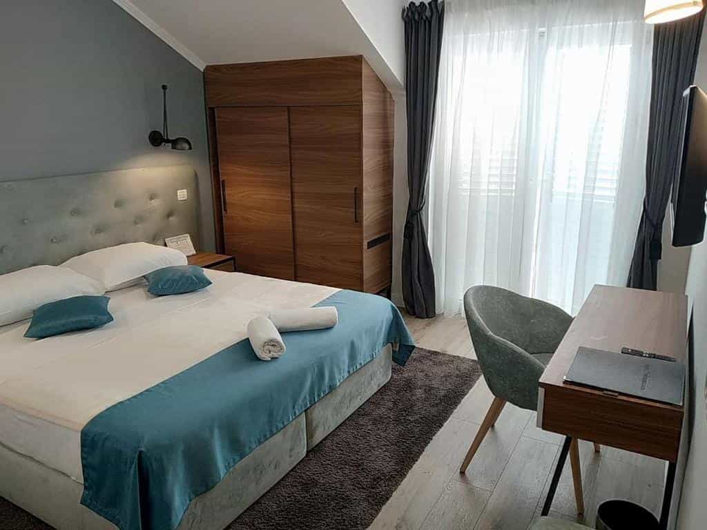 Hotel room interior with large bed, wooden wardrobe, desk, and sheer curtains over a window.