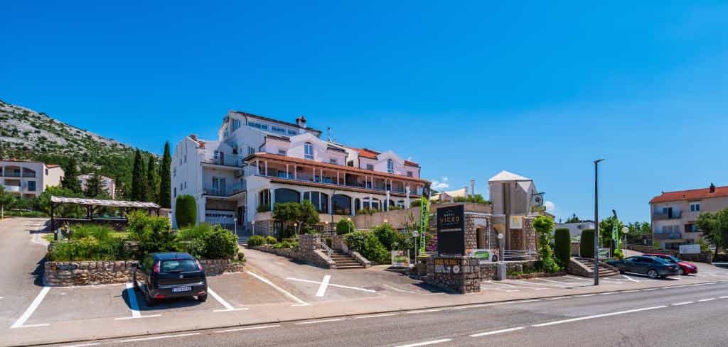 White Hotel Vicko building with parking lot and rocky hillside under clear blue sky.