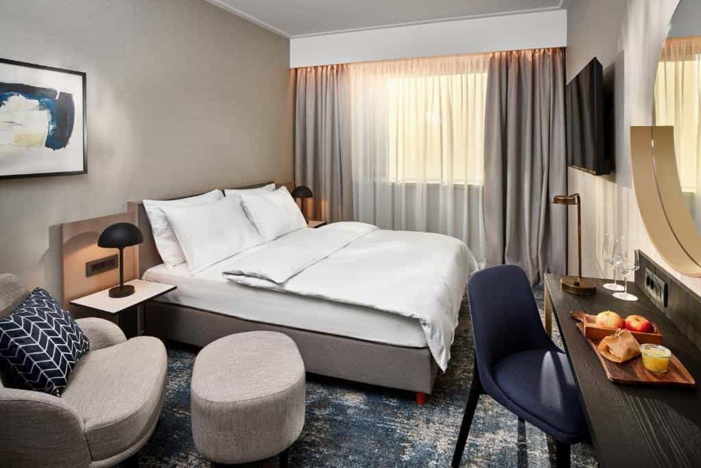 Modern hotel bedroom with white bedding, accent chair, desk, and bright window light.