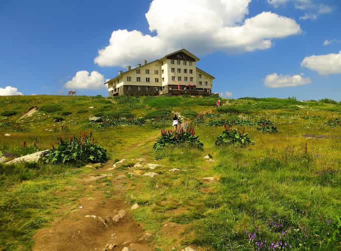 Hikers approaching Rila Lakes Hut on a grassy mountain slope under a blue sky.