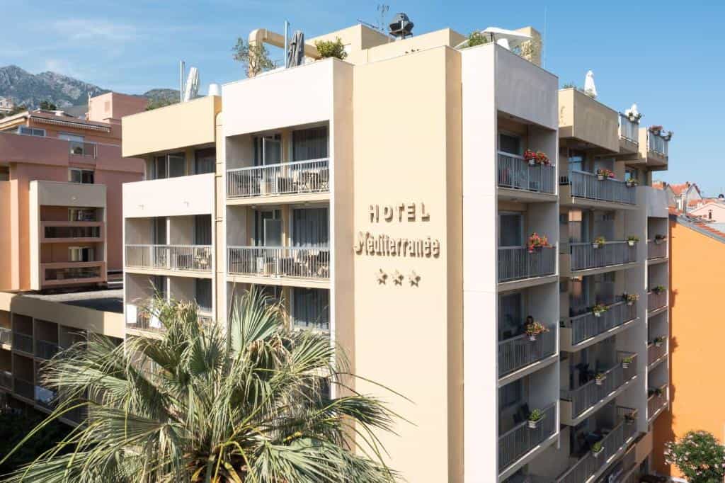 Hotel Mediterranée building with balconies, palm tree, and mountains in the background.