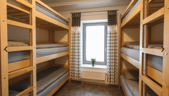 Wooden triple-stacked bunk beds in a hostel room with a bright window and plaid curtains.