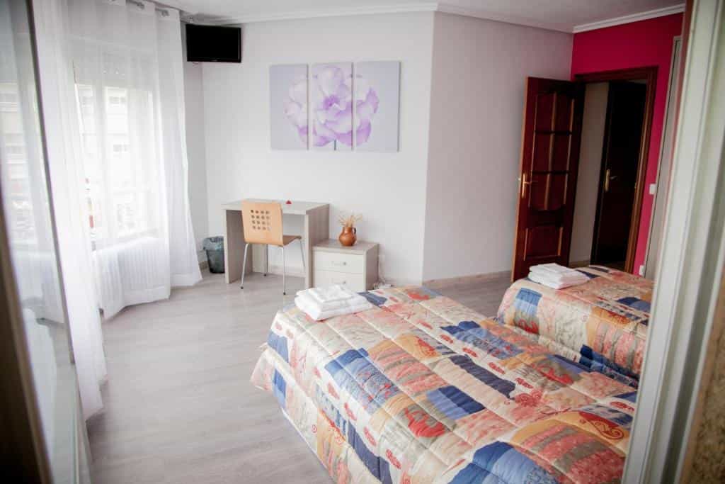 Twin beds with patterned quilts in a bright room with white sheer curtains and a desk.