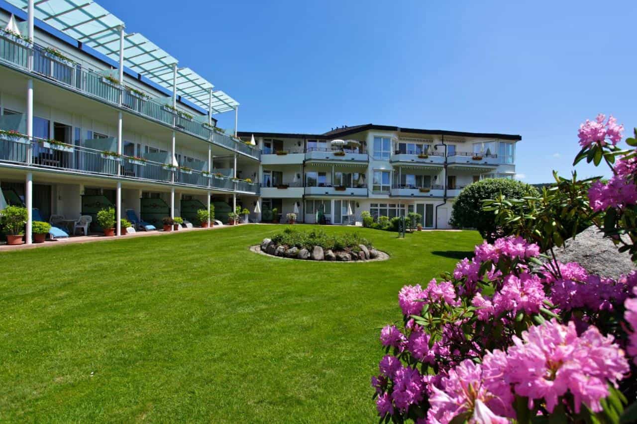 Parkhotel Flora am Schluchsee building with balconies overlooking a green lawn and pink rhododendrons.