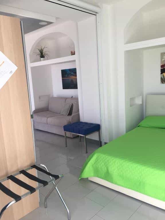 Brightly lit hotel room with white walls, a bed with a lime green cover, and a view into a sitting area.