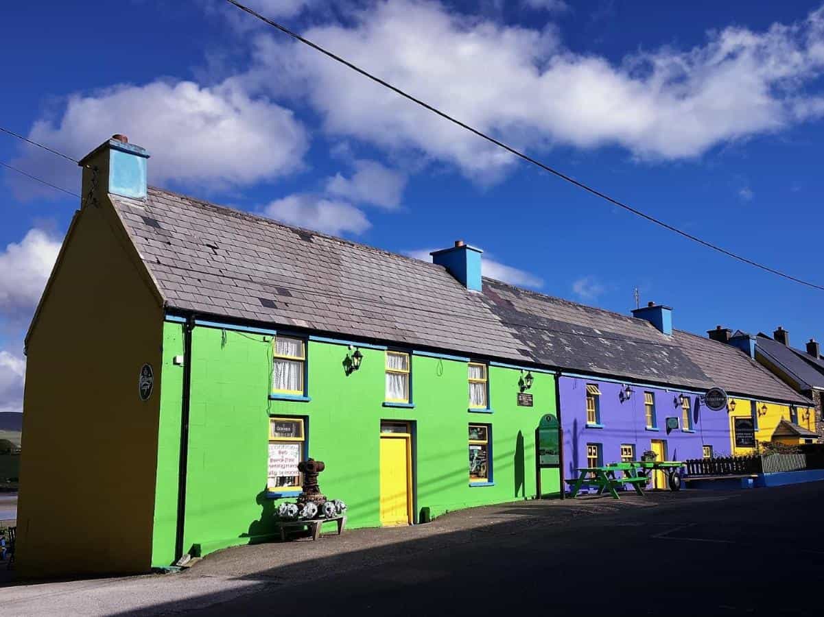 Row of brightly painted guesthouse buildings with slate roofs under a blue, cloudy sky.