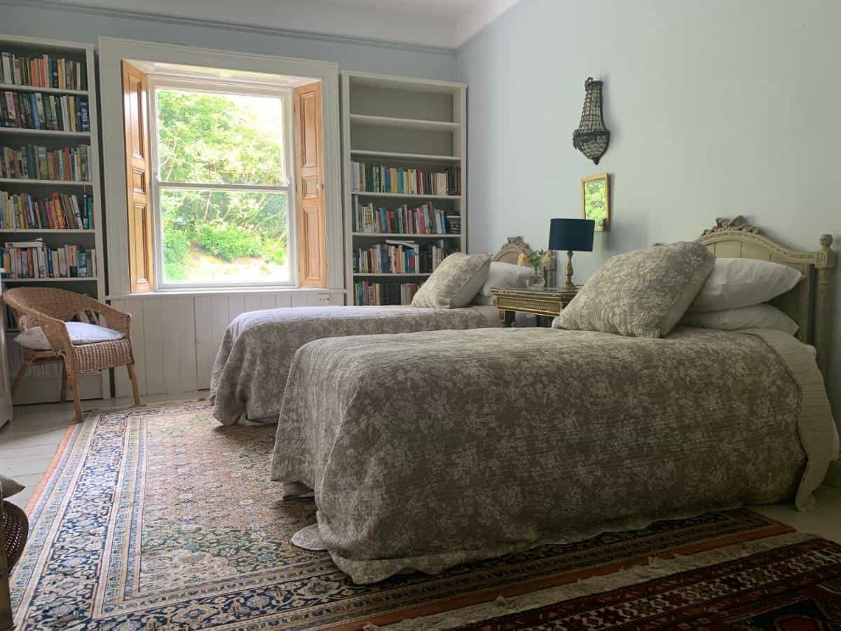 Twin beds in a bright room with built-in bookshelves and a window view of greenery.