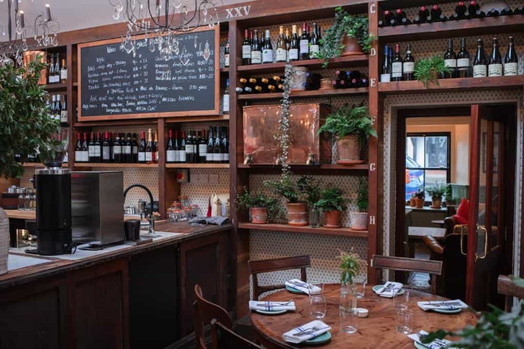 Interior of a restaurant or bar with a wooden counter, wine shelves, and a set dining table.