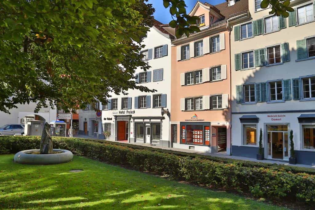 Hotel Freieck facade next to shops, viewed across a sunny lawn with a stone fountain.