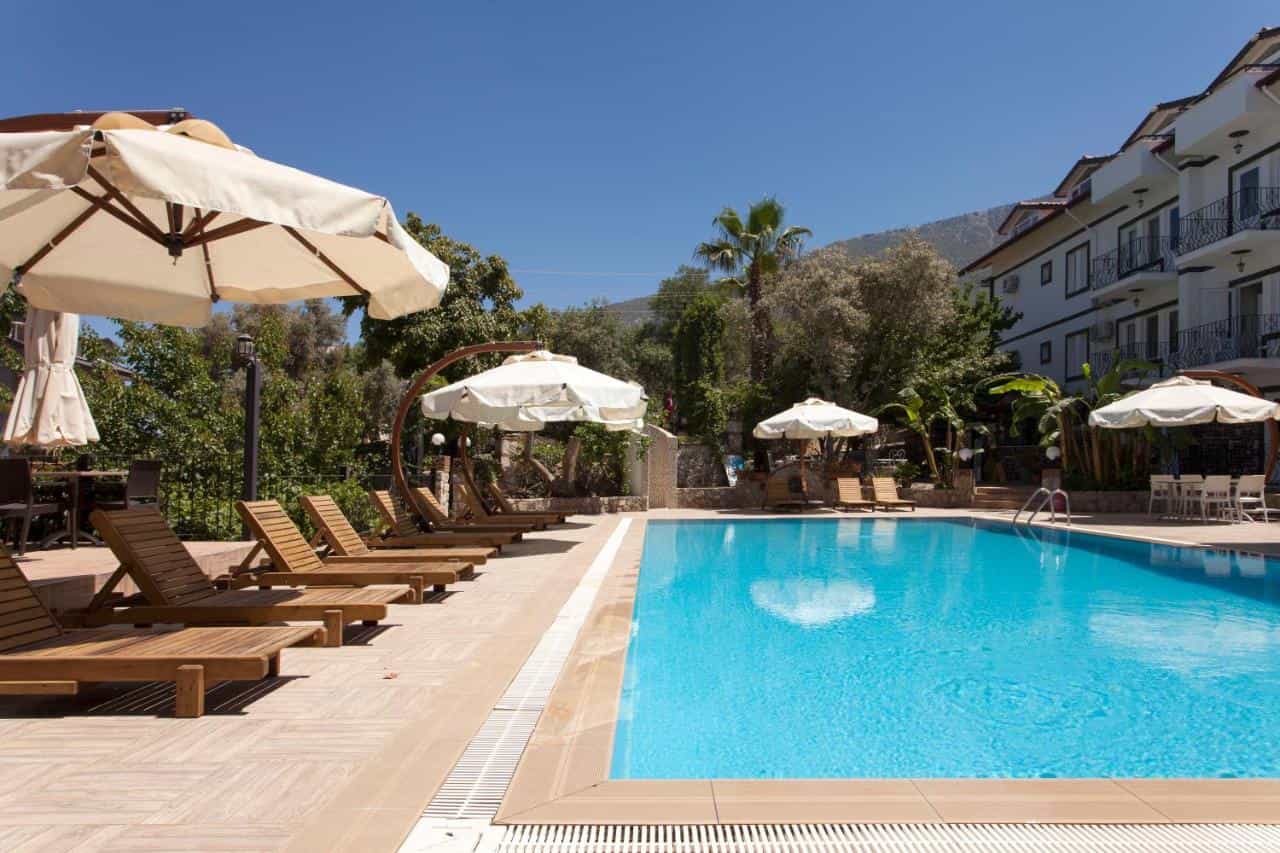 Swimming pool with wooden lounge chairs and umbrellas next to a white hotel building and lush greenery.