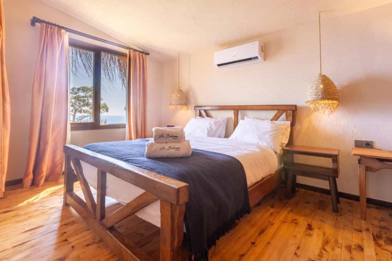 Bedroom with wooden bed, white linens, and view of sea and tree through window