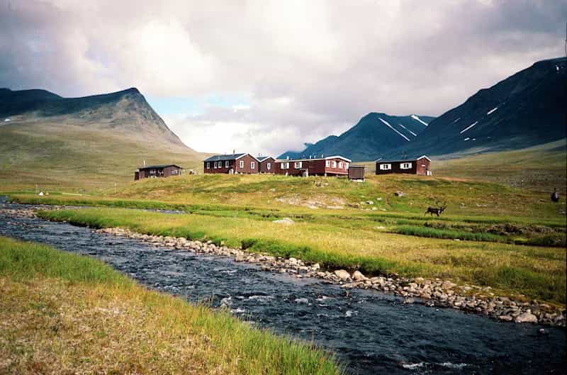 Red wooden cabins by a mountain stream in Sälka, Sweden, with reindeer grazing in the valley.