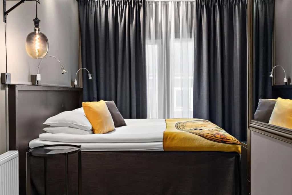 Hotel room bed with white linens, yellow pillows, and dark curtains in front of a window.