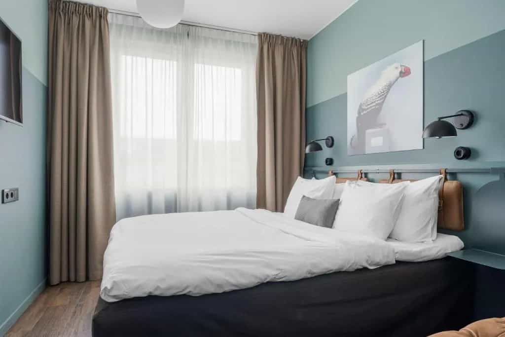 Bedroom with white bedding, blue-green walls, and a large window with sheer and brown curtains.
