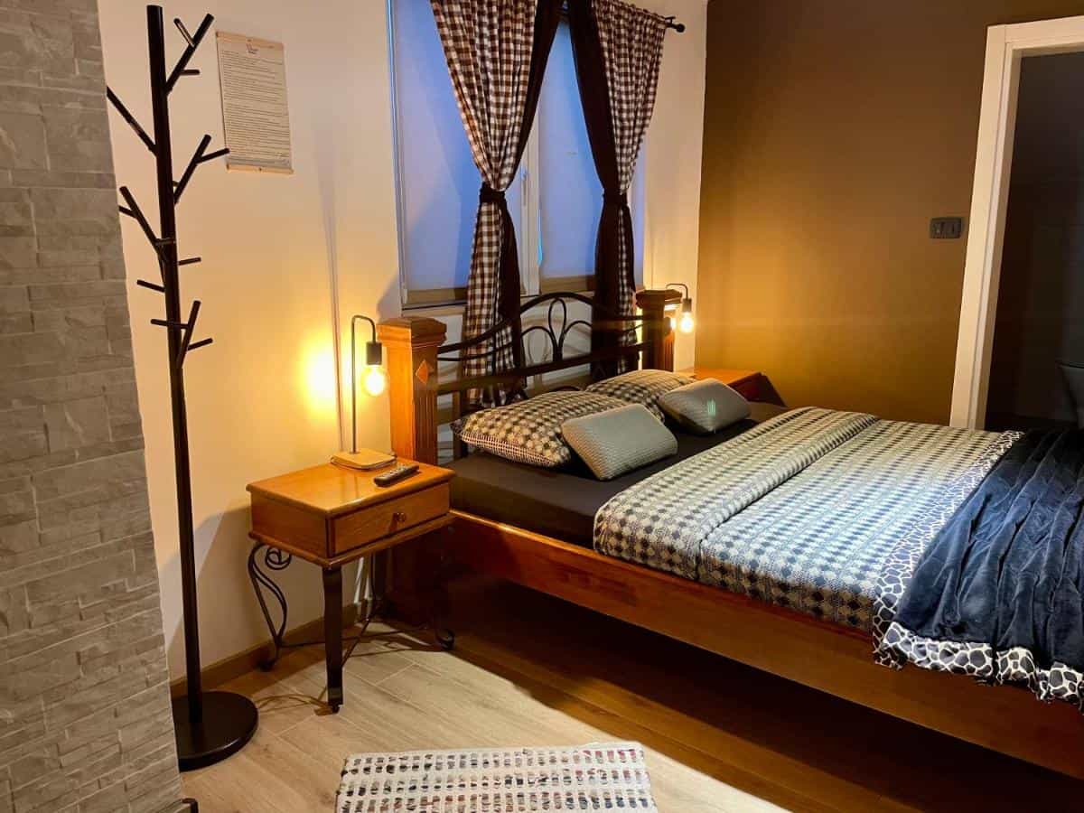 Cozy guest room with wooden bed, patterned bedding, and warm lighting near a window with checkered curtains.