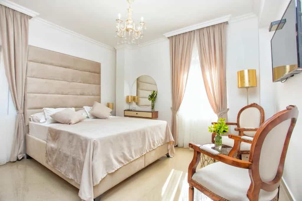 Hotel Admiral room with beige bedding, tufted headboard, chandelier, and two wooden accent chairs.