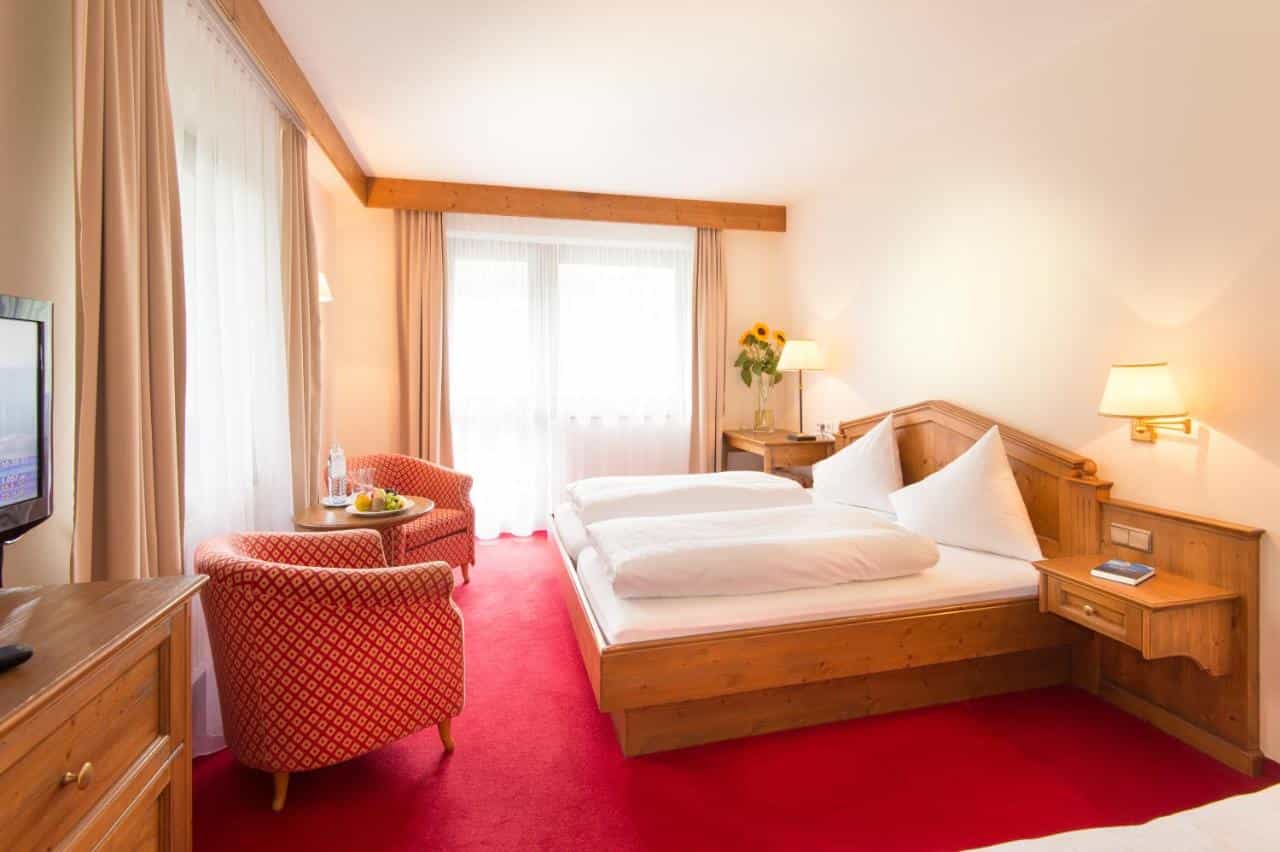Hotel room with twin beds, red carpet, wooden furniture, and bright window light.
