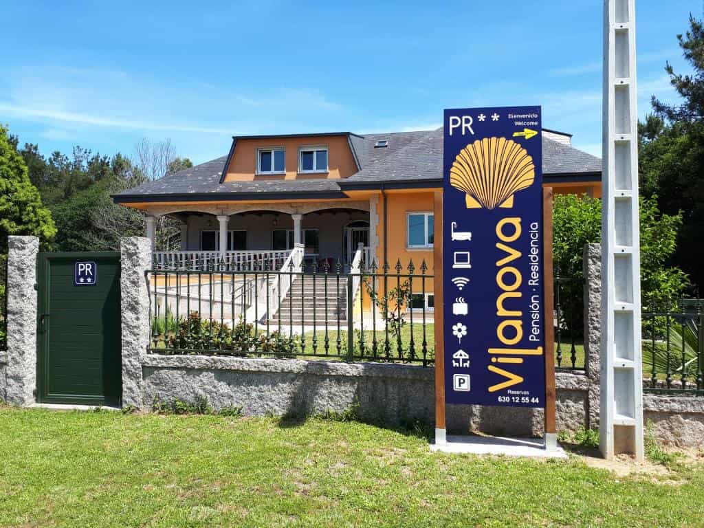 Pensión Vilanova sign with scallop shell logo in front of orange house and green lawn.