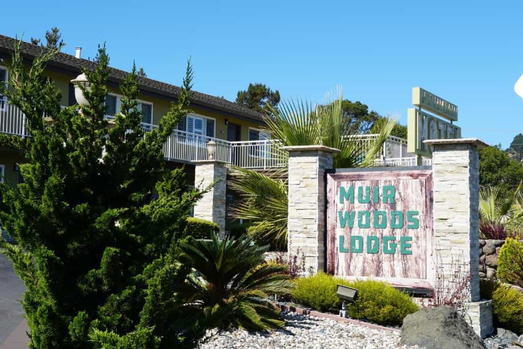Muir Woods Lodge sign with stone pillars, lush greenery, and a two-story building under a clear blue sky.