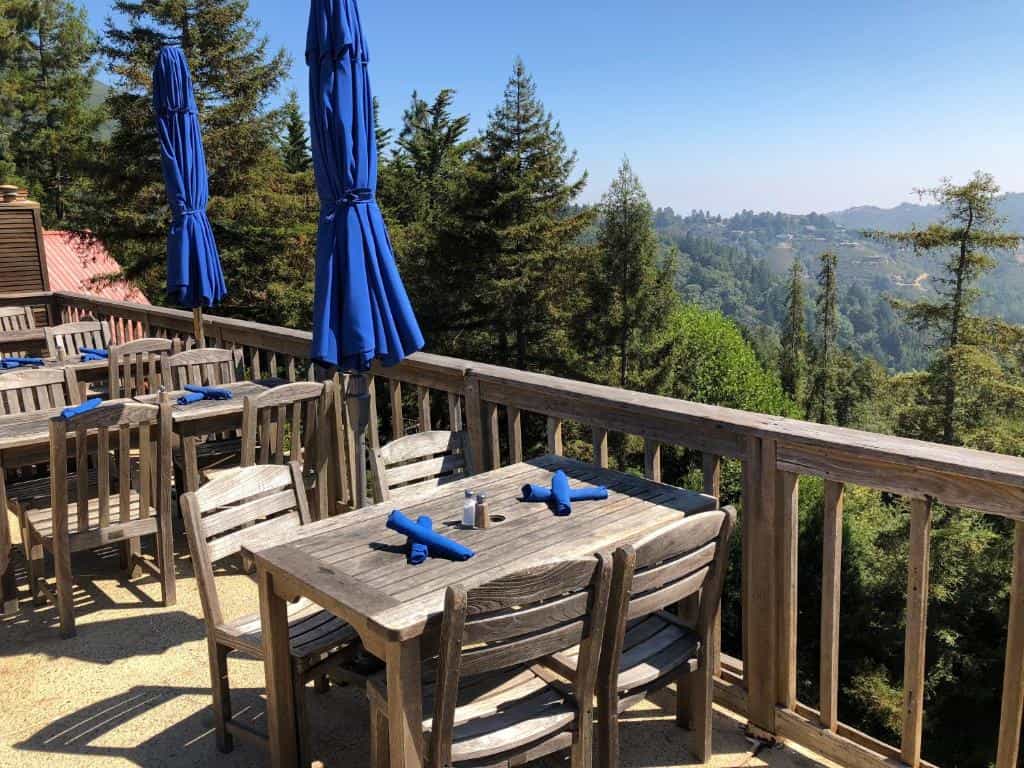 Outdoor dining deck with wooden tables, chairs, and blue umbrellas overlooking forested hills.