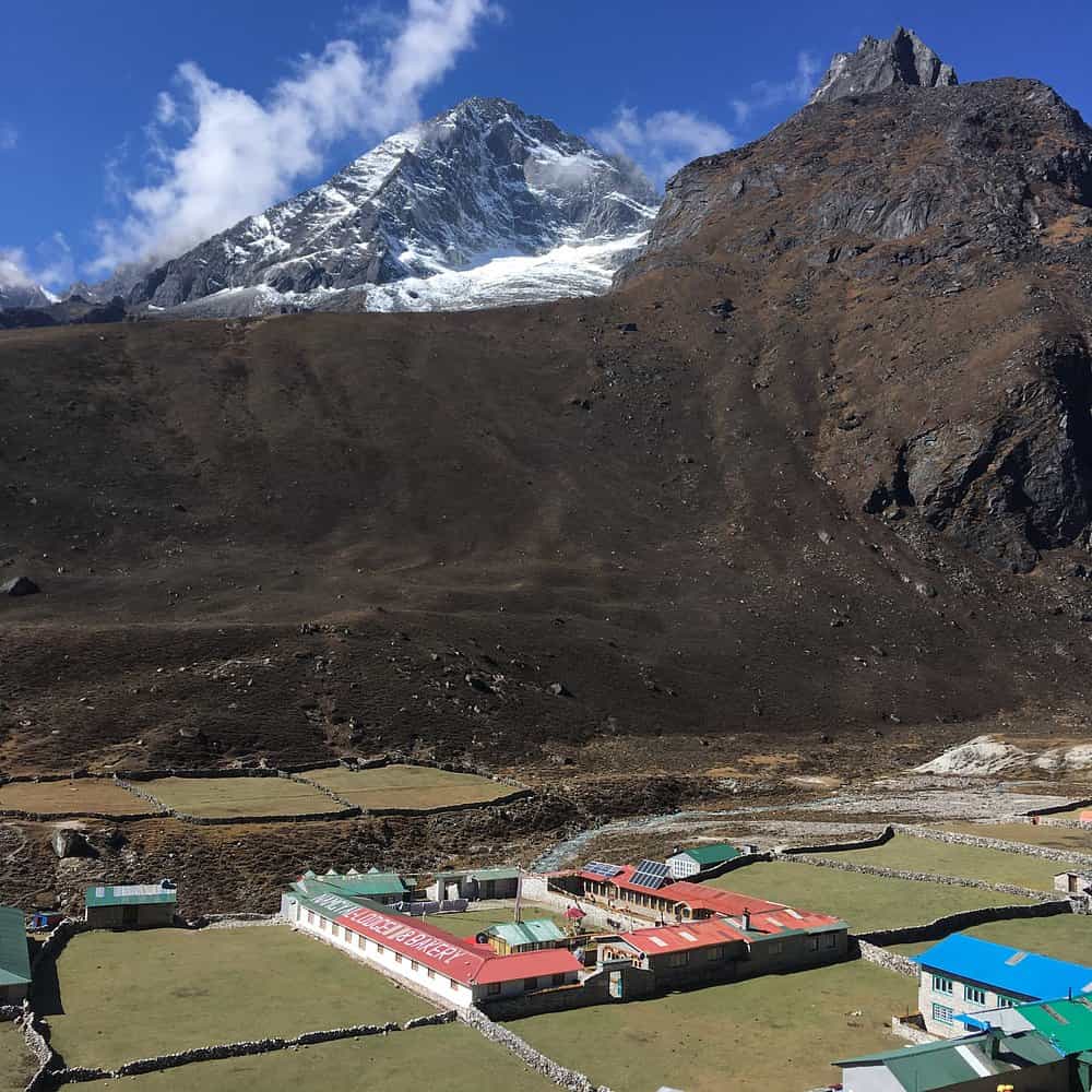 Machhermo Lodge and Bakery buildings in a green valley below massive rocky, snow-capped Himalayan mountains.