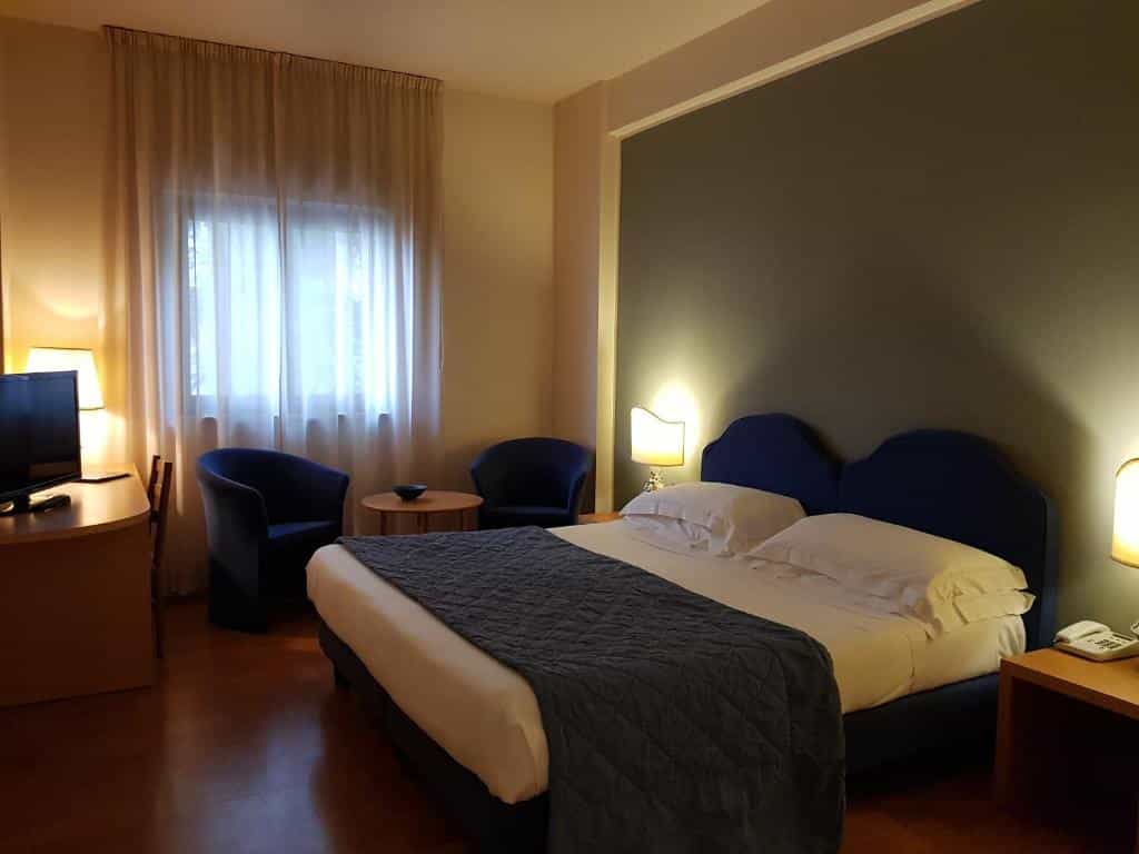Hotel room with double bed, blue accent chairs, desk, and window with sheer curtains.