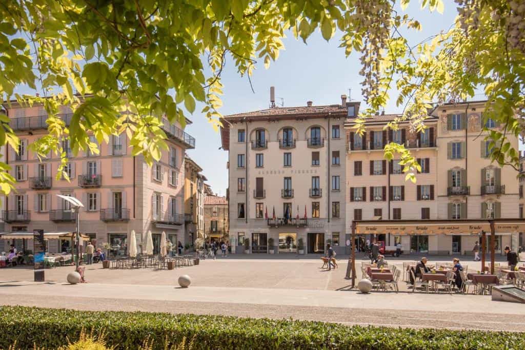 Albergo Firenze hotel facade on sunny piazza with outdoor cafe seating framed by green leaves.