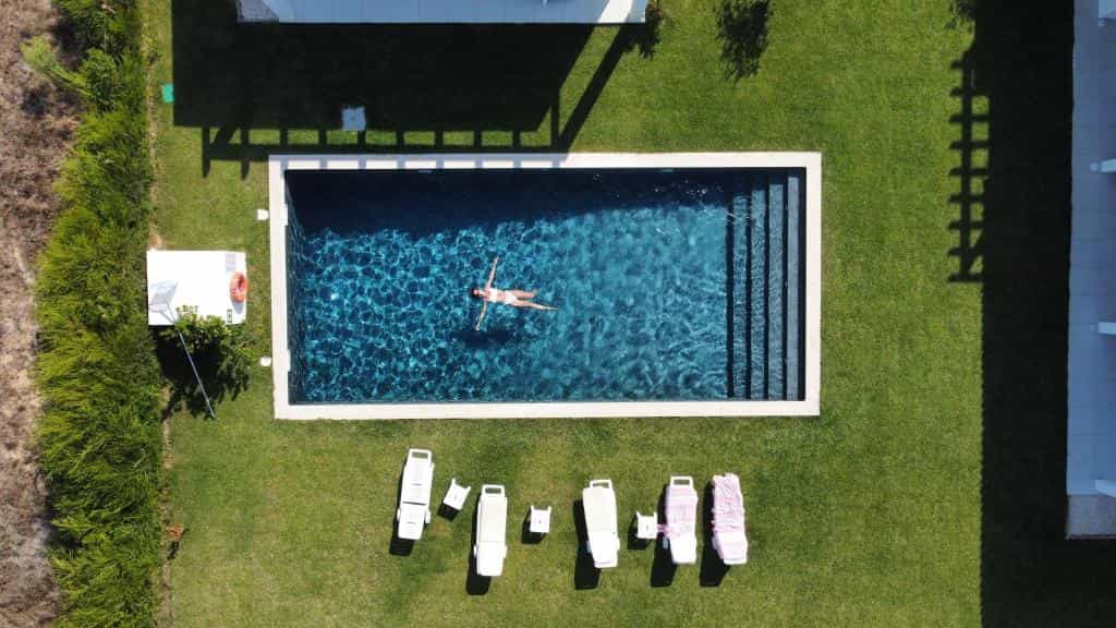 Person floating in a rectangular swimming pool surrounded by a green lawn with lounge chairs.