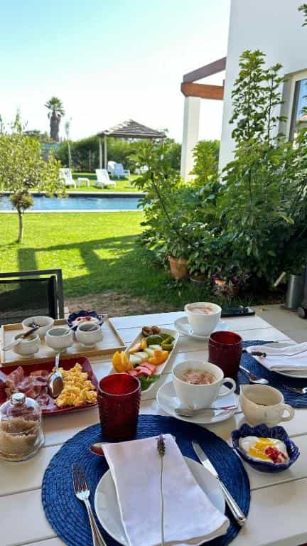 Breakfast spread with coffee, fruit, and eggs on a patio table overlooking a swimming pool and garden.