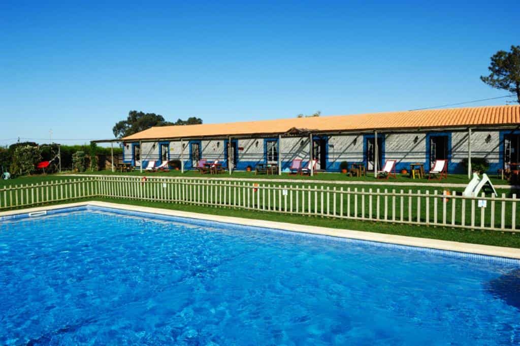 Swimming pool with blue water in front of a long, single-story building with a tiled roof and lounge chairs.
