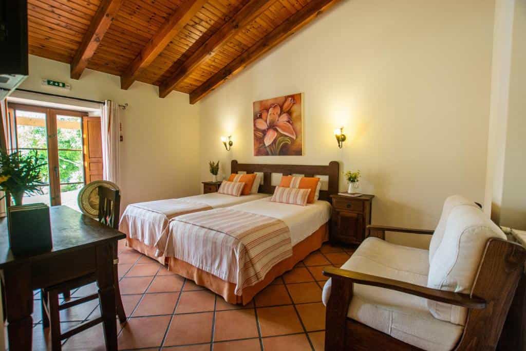 Hotel bedroom with two twin beds, wooden ceiling beams, tiled floors, and a small sitting area.