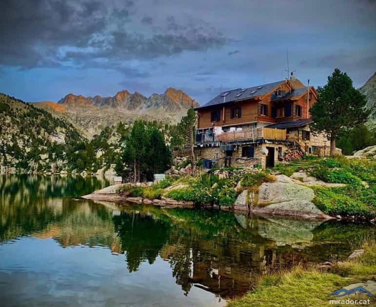 Refugi Josep Maria Blanc mountain hut by a lake with surrounding peaks and pine trees.