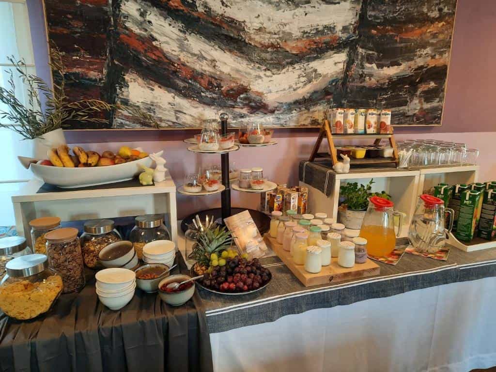Breakfast buffet spread with fruit, yogurt, cereal, and juice on a table in front of an abstract painting.