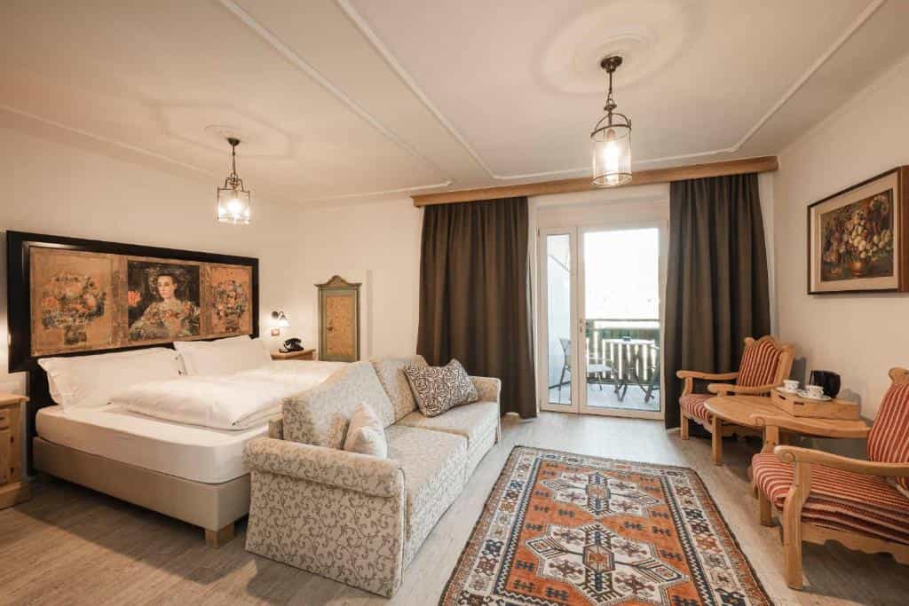 Hotel room with a double bed, patterned sofa, wooden chairs, and a balcony door.