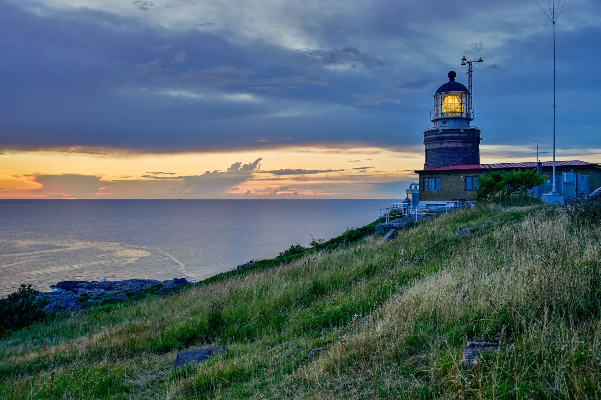 Illuminated lighthouse on grassy cliff overlooking the sea at sunset with dark blue sky.