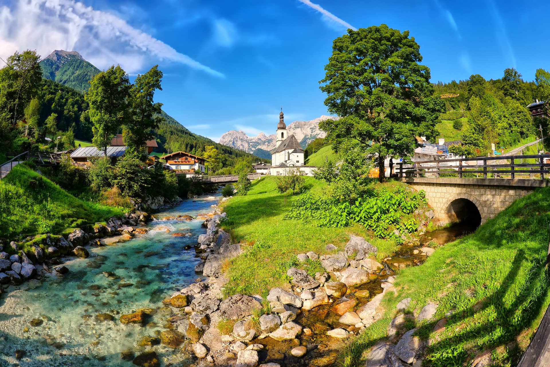 Alpine village with clear stream, church, and mountains under a bright blue sky.