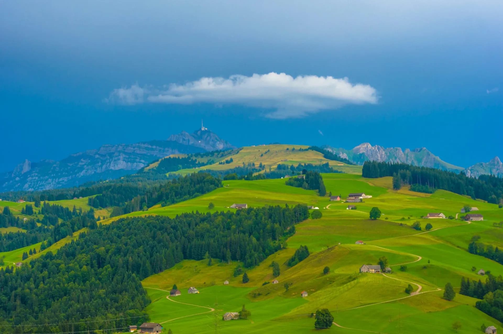 Rolling green hills and forests under stormy blue sky with distant Swiss mountains