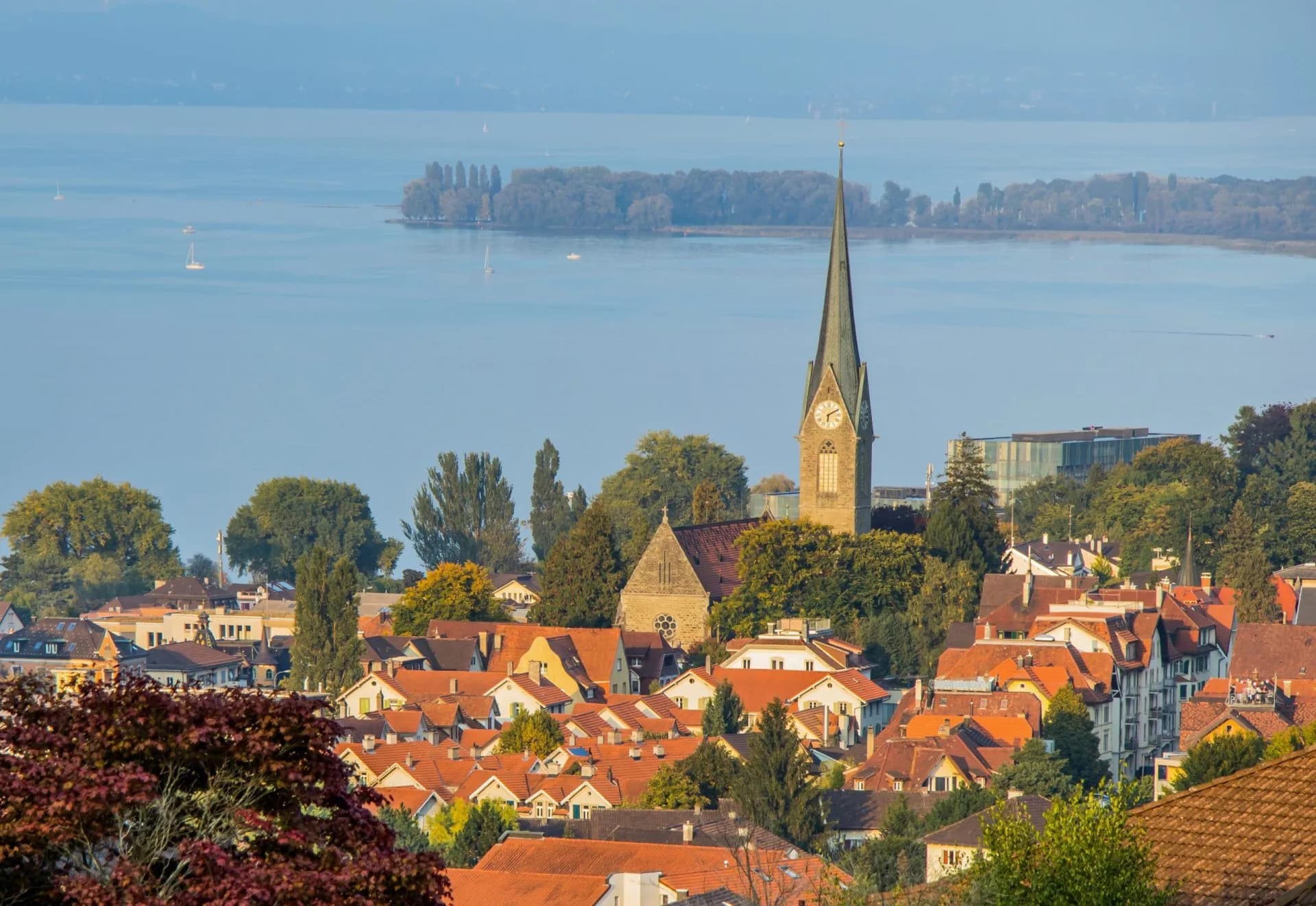 Town with terracotta roofs and church spire overlooking Lake Constance with sailboats.