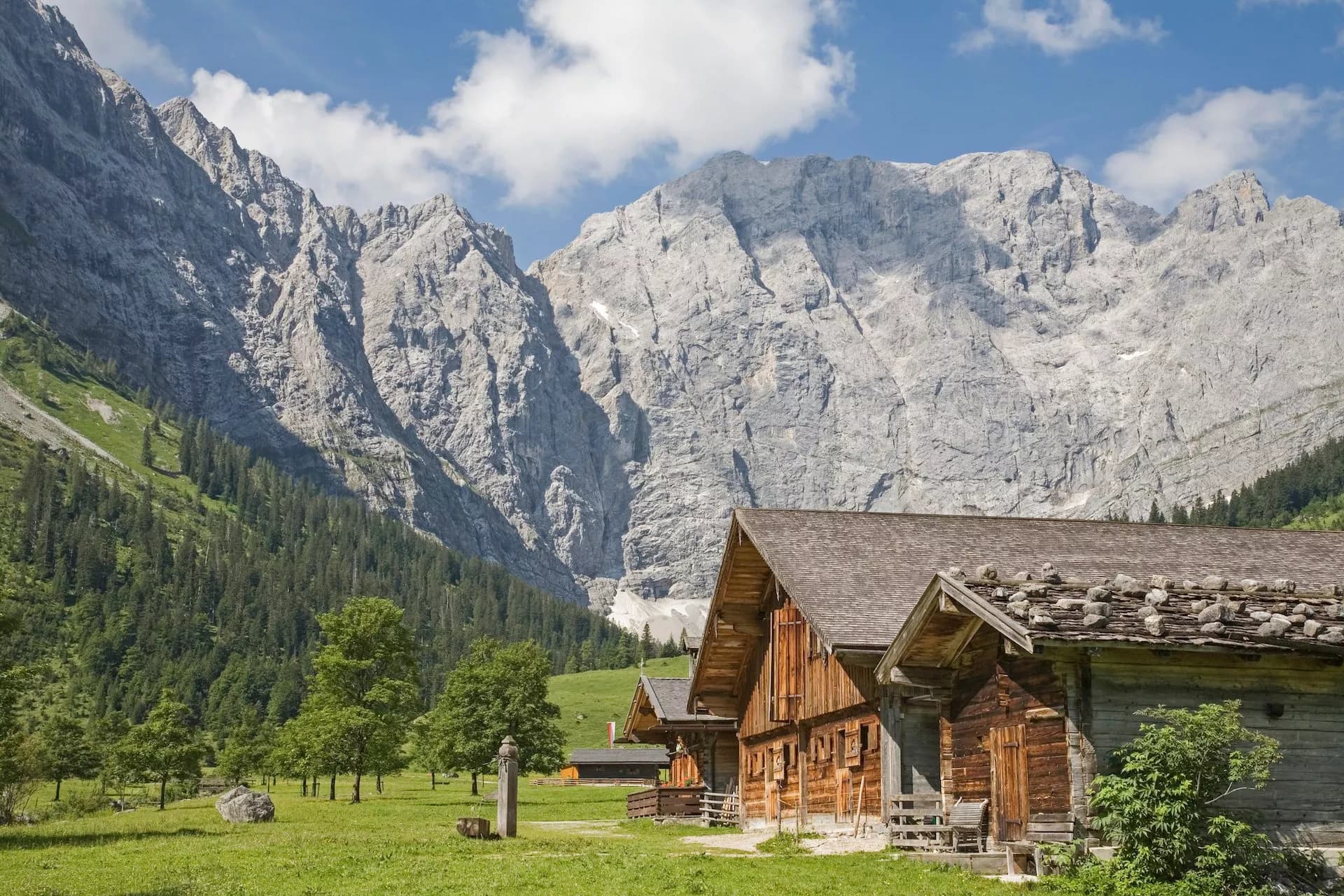 Wooden alpine village huts in a green meadow below massive gray mountains under a blue sky.