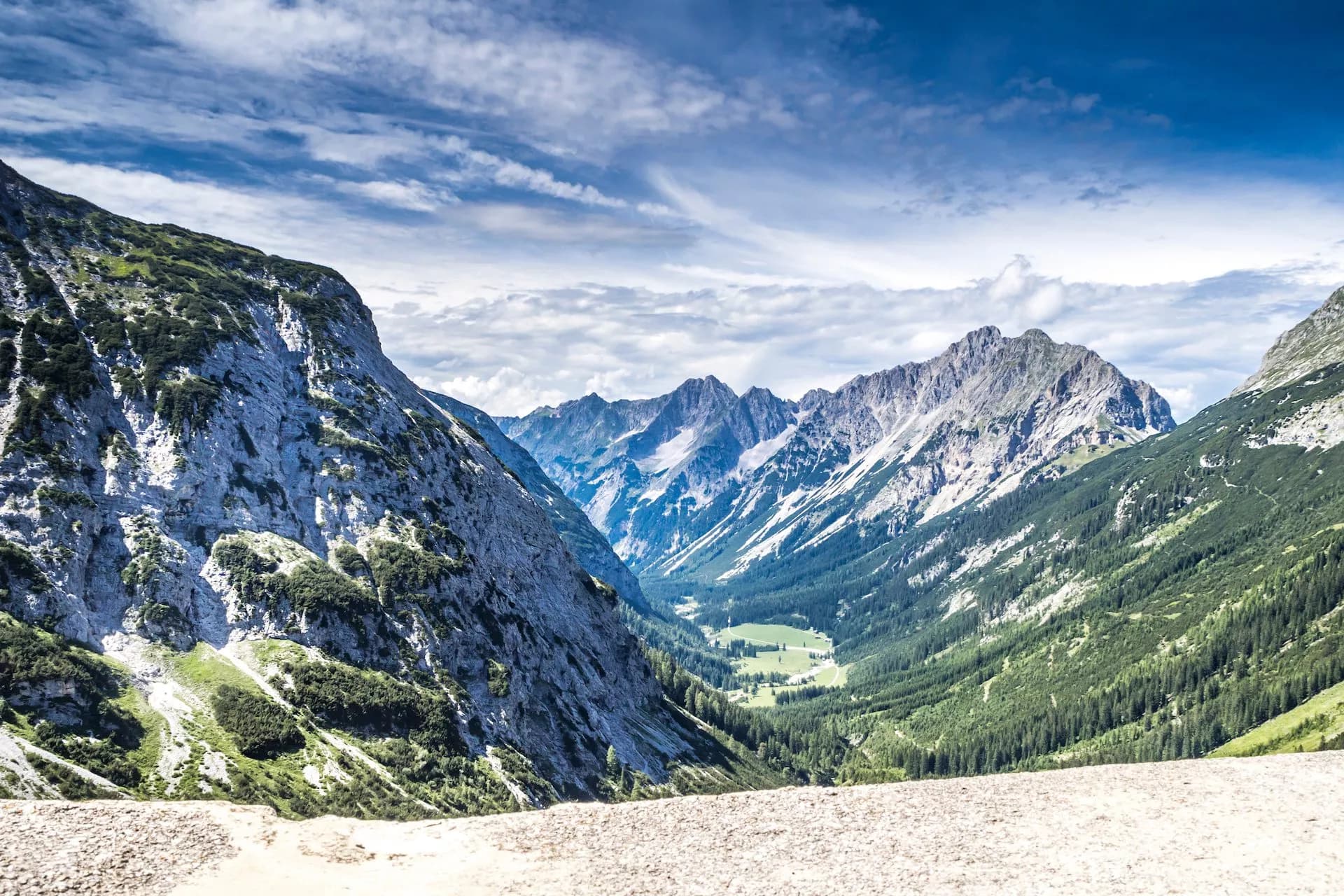 Karwendel Valley with steep, rocky mountains, green forests, and a valley floor under a cloudy blue sky.