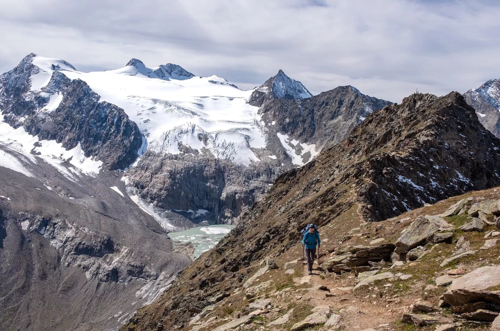 Hiker with poles on rocky trail below massive snow-capped glacier and glacial lake