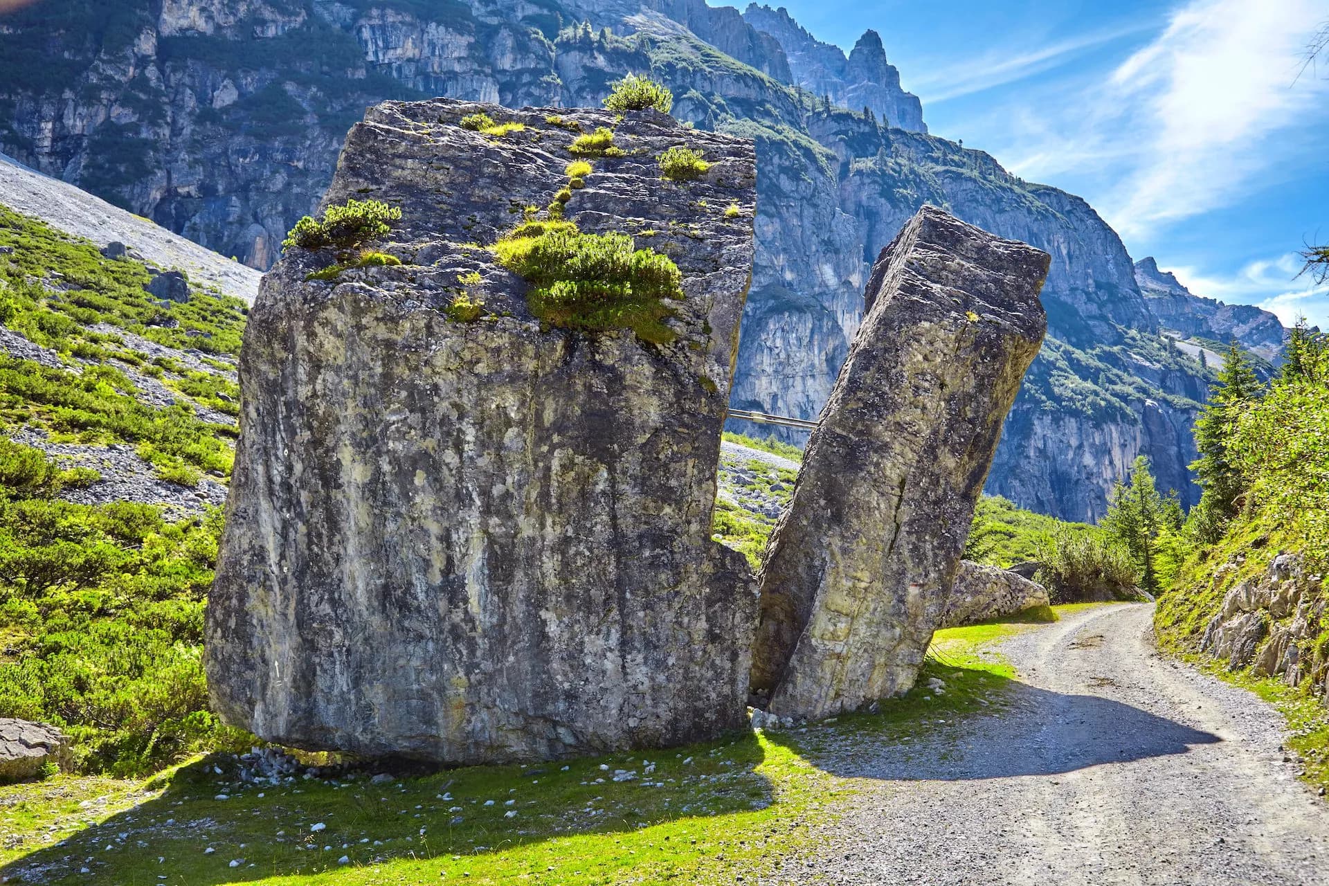 Large split rock formation next to gravel path with steep, green mountains under blue sky.