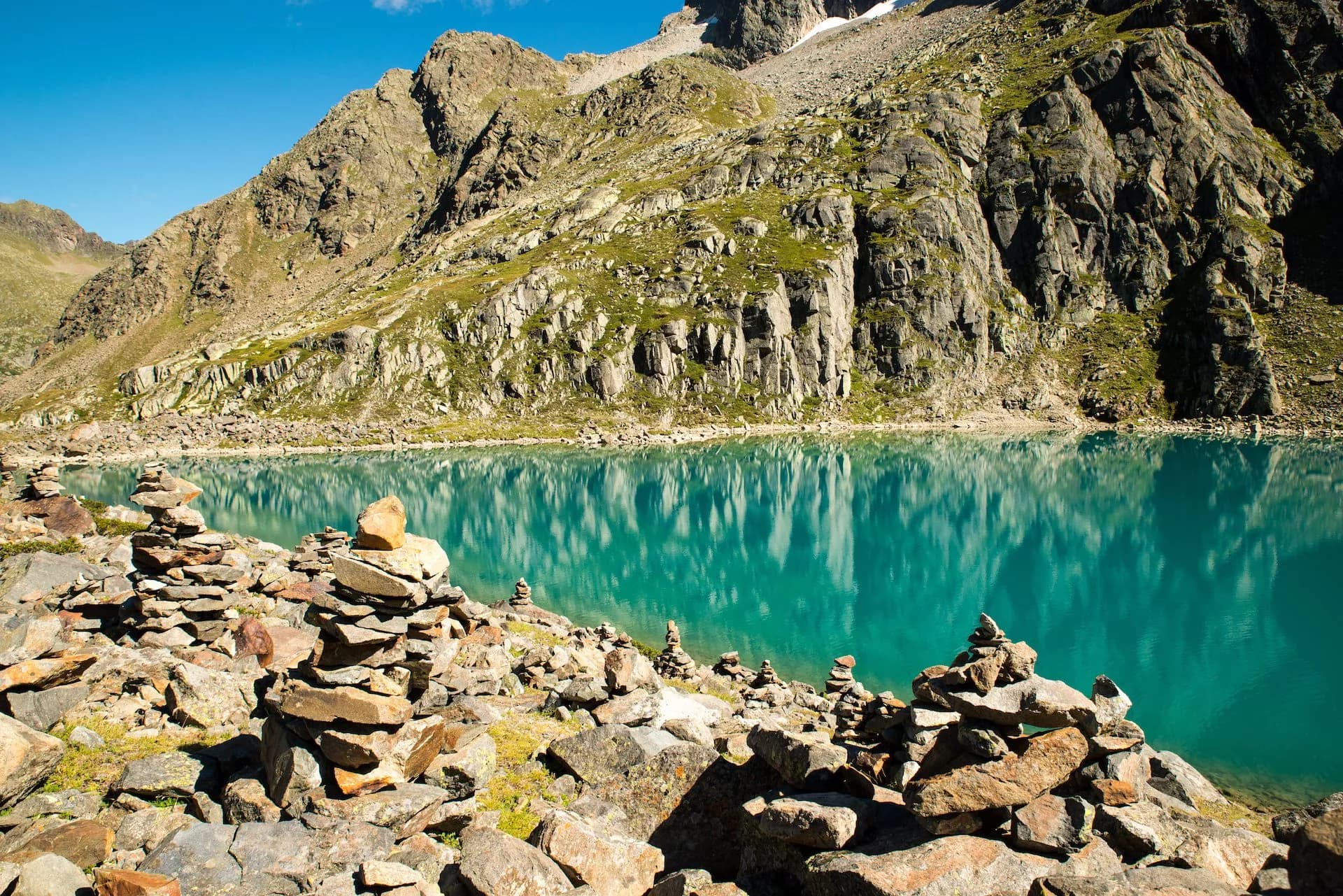 Rock cairns on shore of turquoise alpine lake reflecting steep, rocky mountains under blue sky.