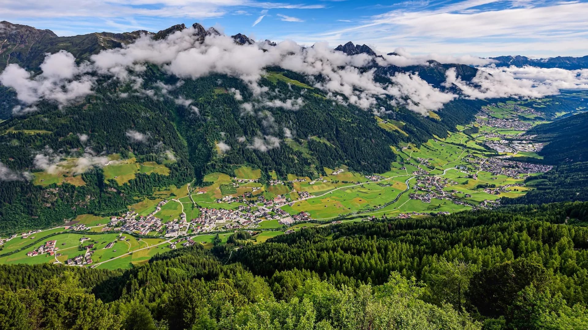 Alpine valley with green meadows, dense forest, and village nestled below cloud-covered mountains in Stubai Valley.