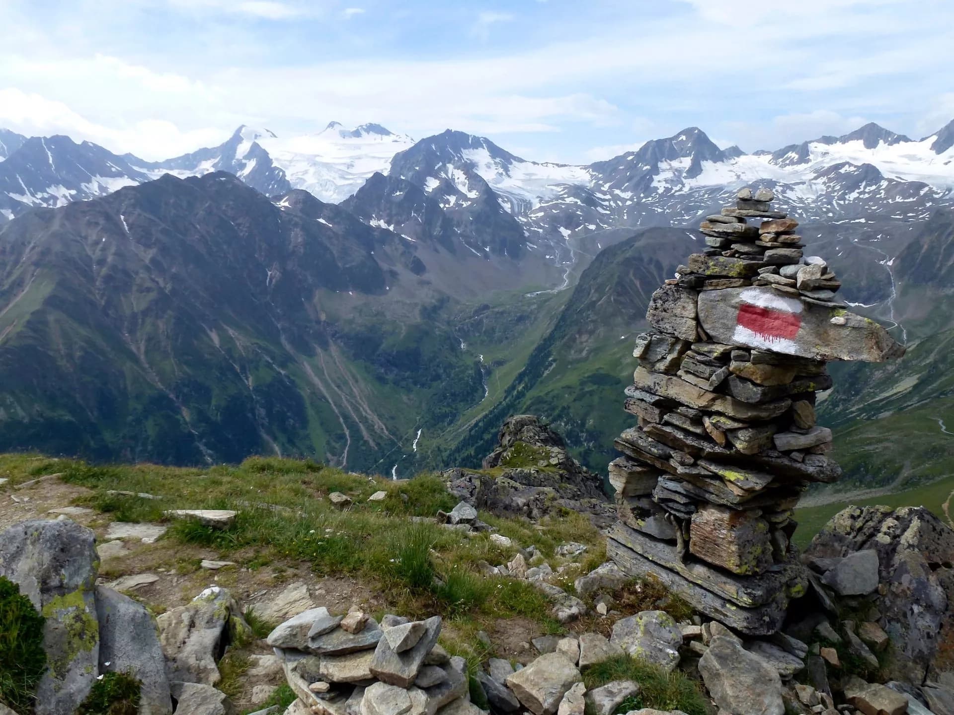 Hiking cairn with red and white trail marker overlooking snowy alpine peaks and green valley