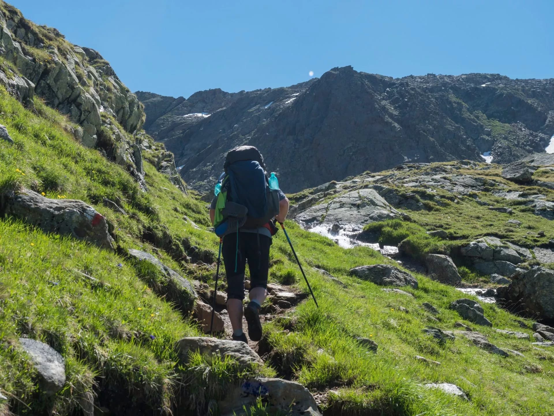 Lonely man hiker with heavy backpack at Stubai hiking trail, Stubai Hohenweg at green summer alpine mountain valley with spring stream. Tyrol, Austrian Alps